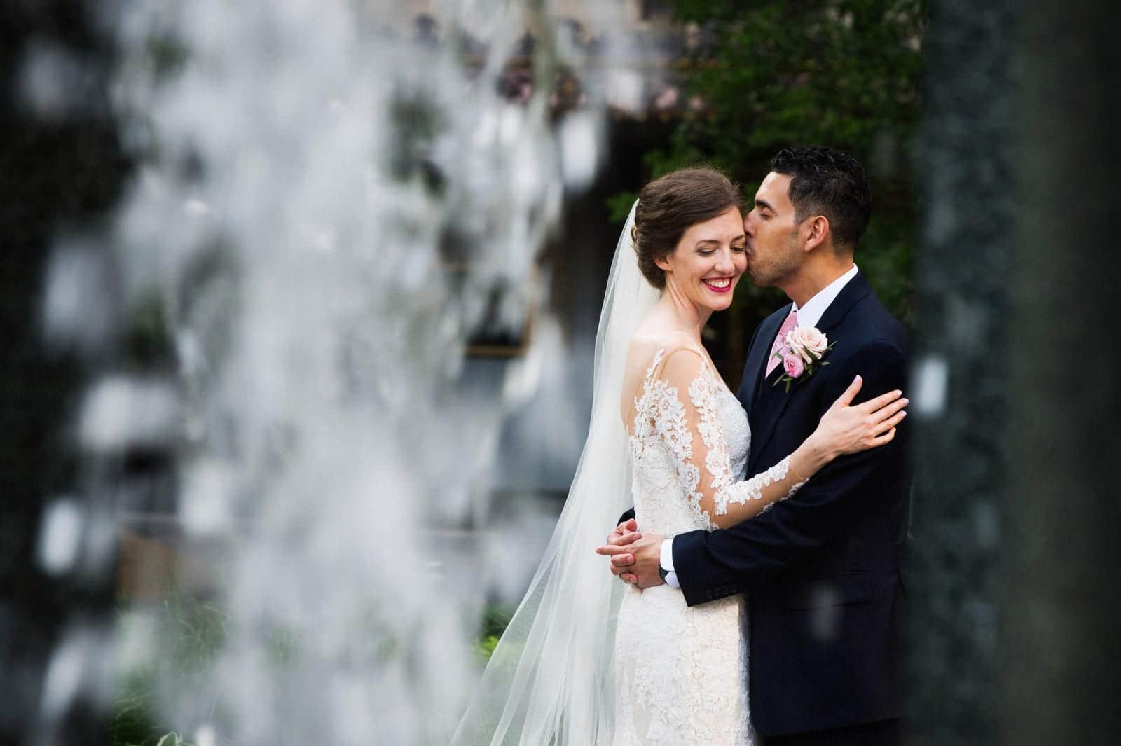 A smiling bride wearing a dress with lace sleeves is kissed on the cheek by her husband as seen through the waterfall across the street from the Omni William Penn.