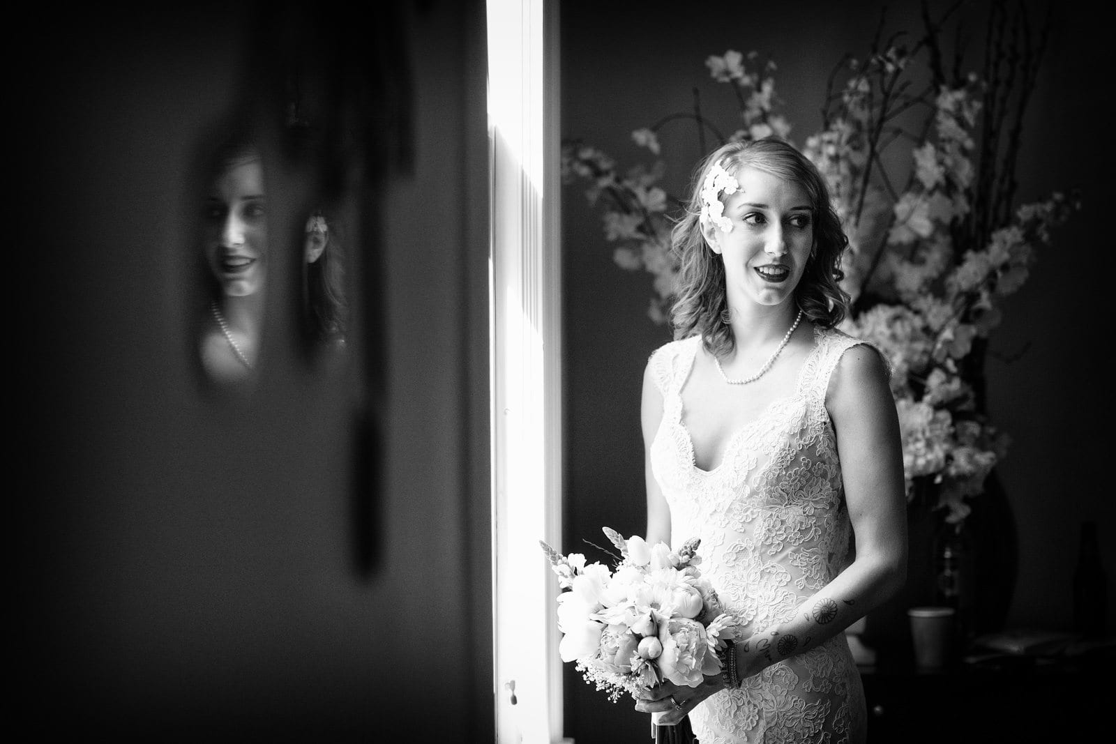 A bride with flowers in her hair stands by a window while her face is reflected in a mirror.