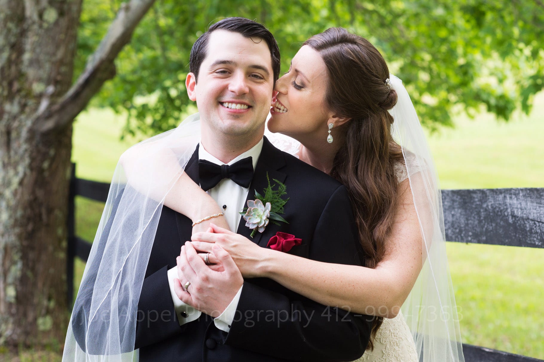 A bride wraps her arms around her tuxedoed groom as she playfully bites his ear lobe during their Chanteclaire Farm Wedding.