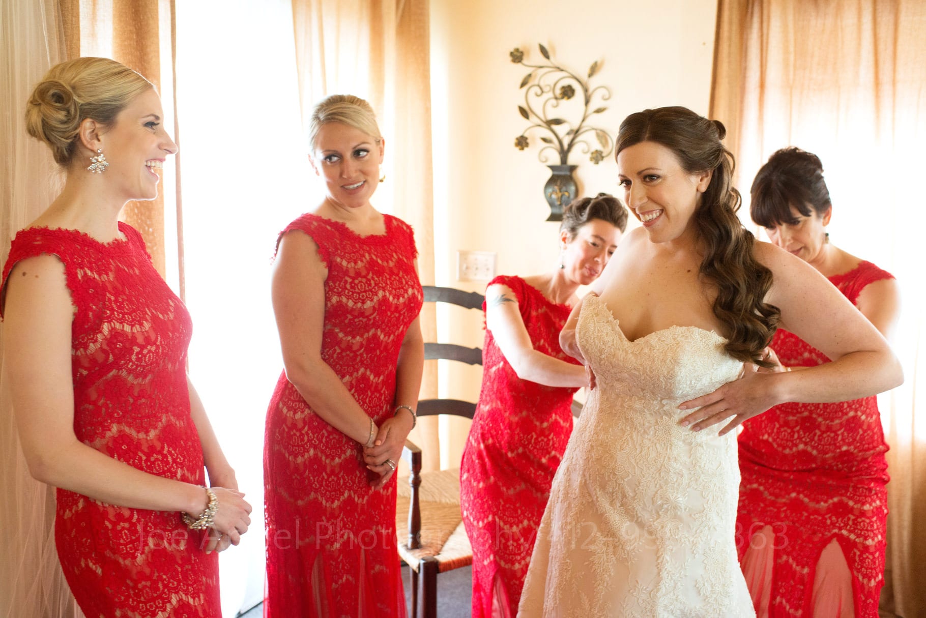 A bride smiles as she is helped into her wedding dress by four bridesmaids all dressed in red lace during their Chanteclaire Farm Wedding.