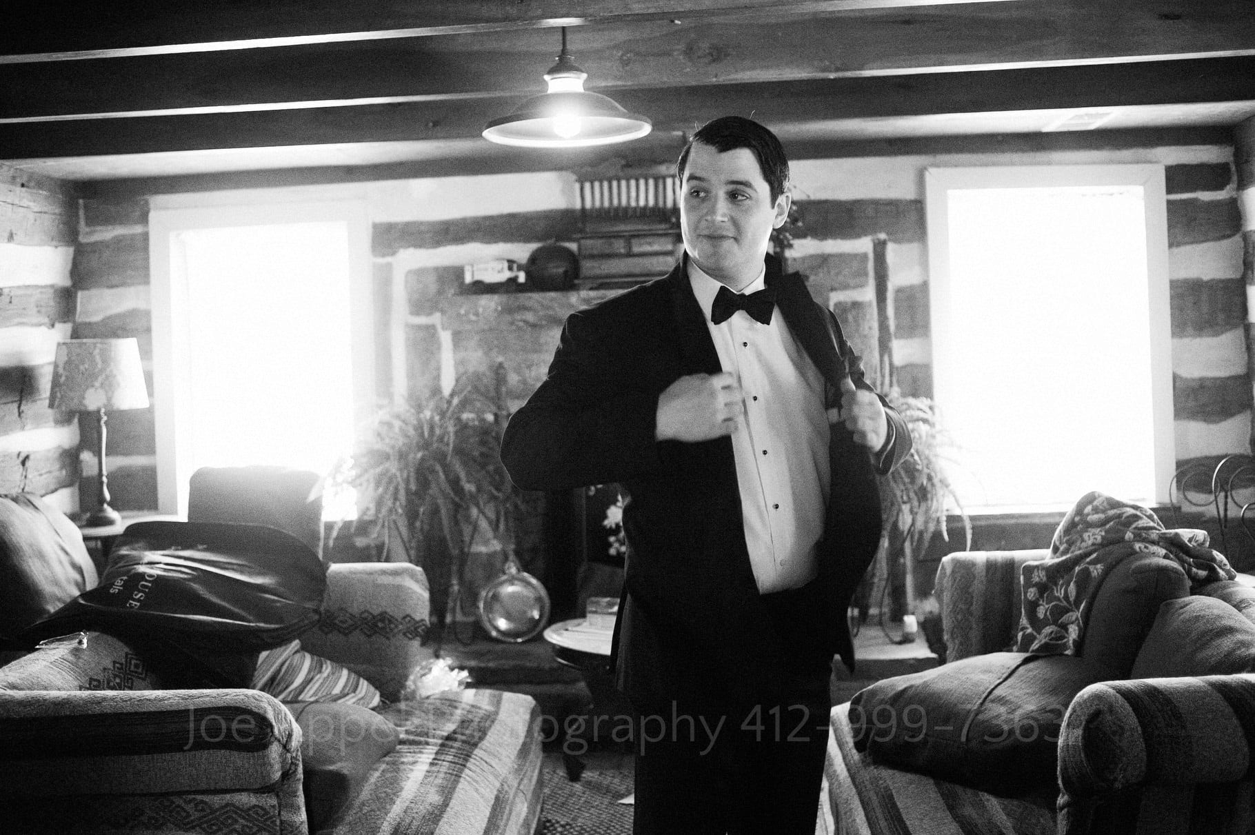a groom holds on to his lapels as he puts on his tuxedo jacket the living room of a log cabin during their Chanteclaire Farm Wedding.