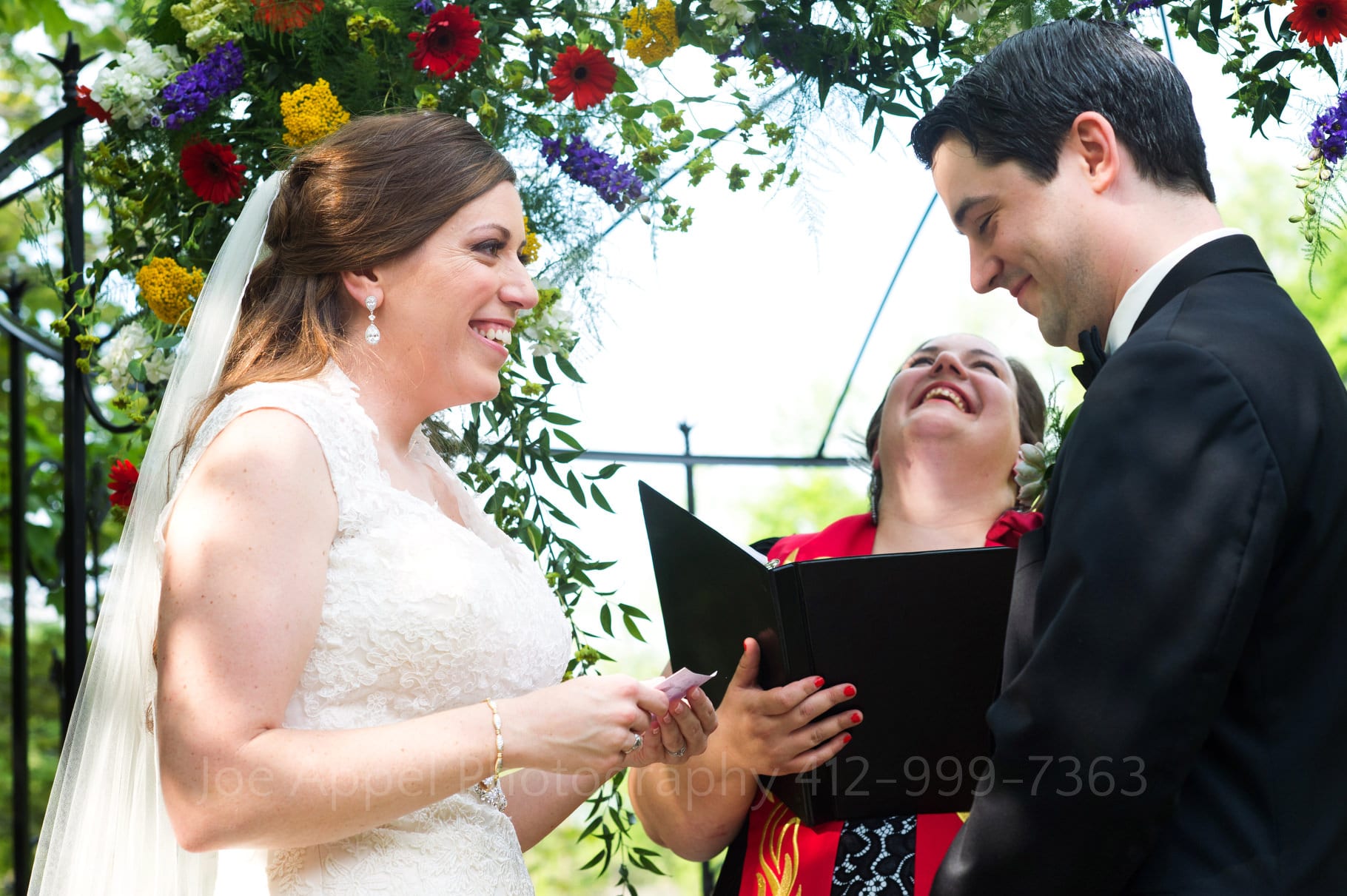 Bride and groom smile as they speak their vows to one another as the officiant rears back her head in laughter during their Chanteclaire Farm Wedding.