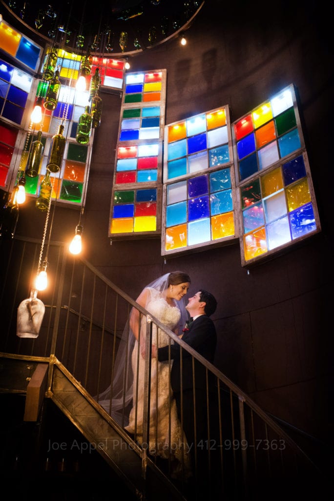 on stairs beneath colored glass windows a bride and groom look at each other during their Chanteclaire Farm Wedding.
