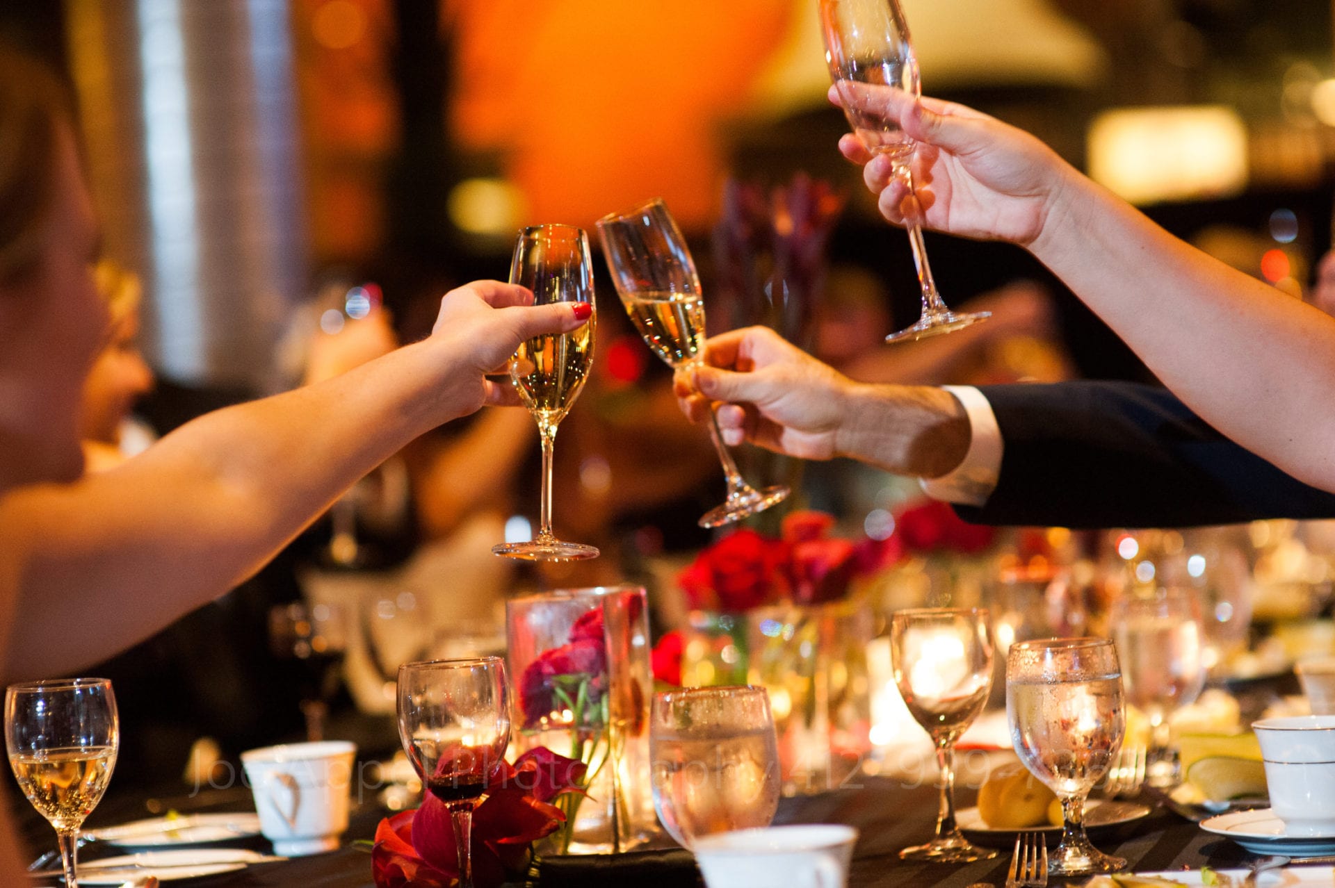 Hands reach out and clink champagne glasses above candle lit tables with red rose centerpieces.
