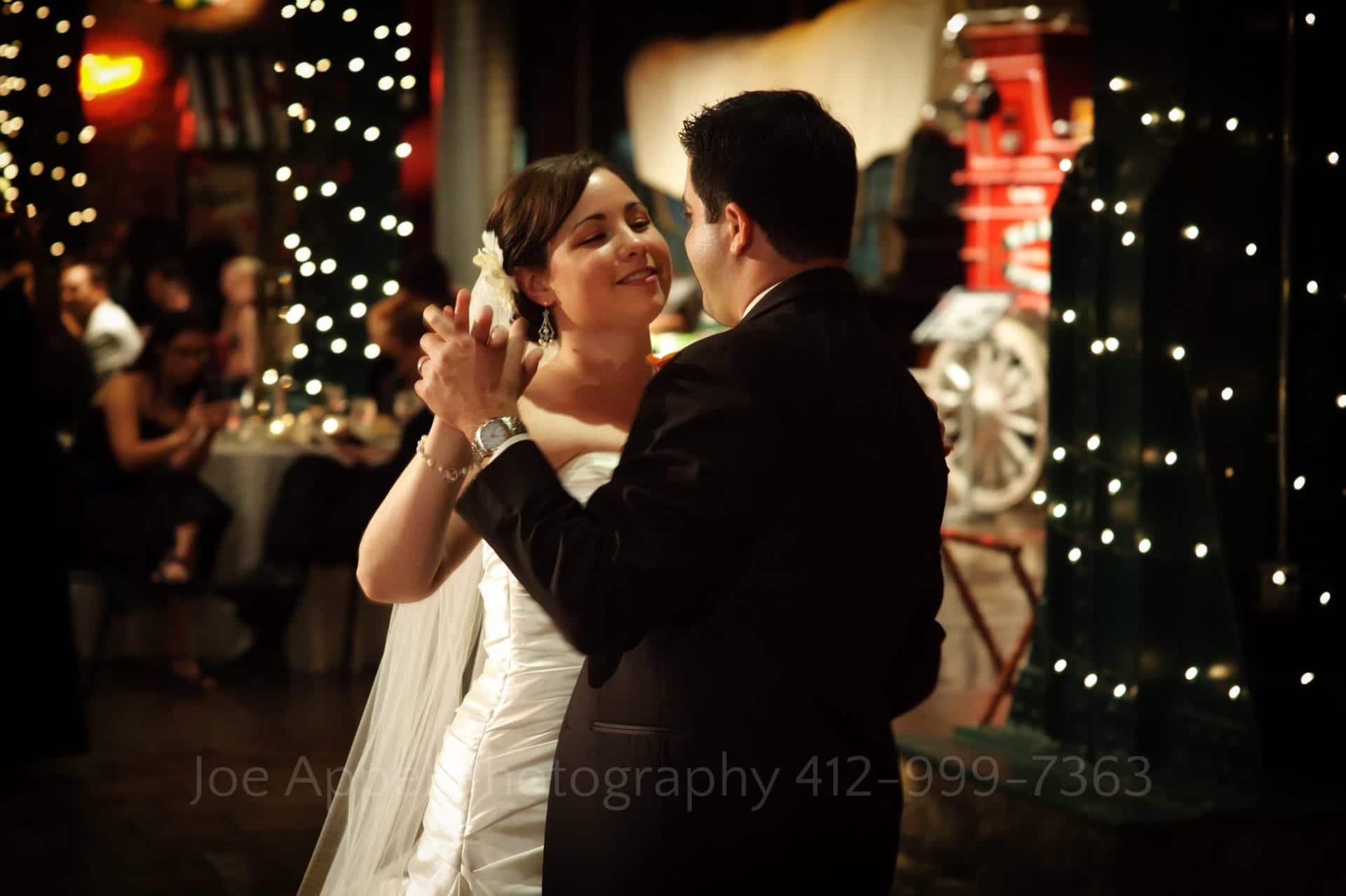 A couple dances at the Great Hall at the Heinz History Center with twinkle lights wrapped around the beams behind them.