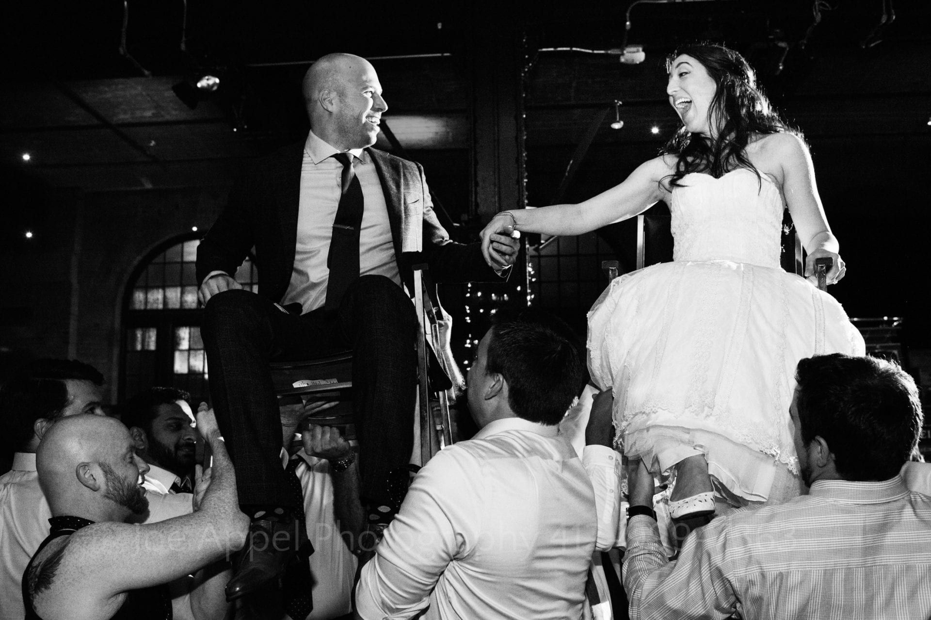 Black and white photo of a bride and groom holding hands as they're lifted up on chairs during their wedding reception.