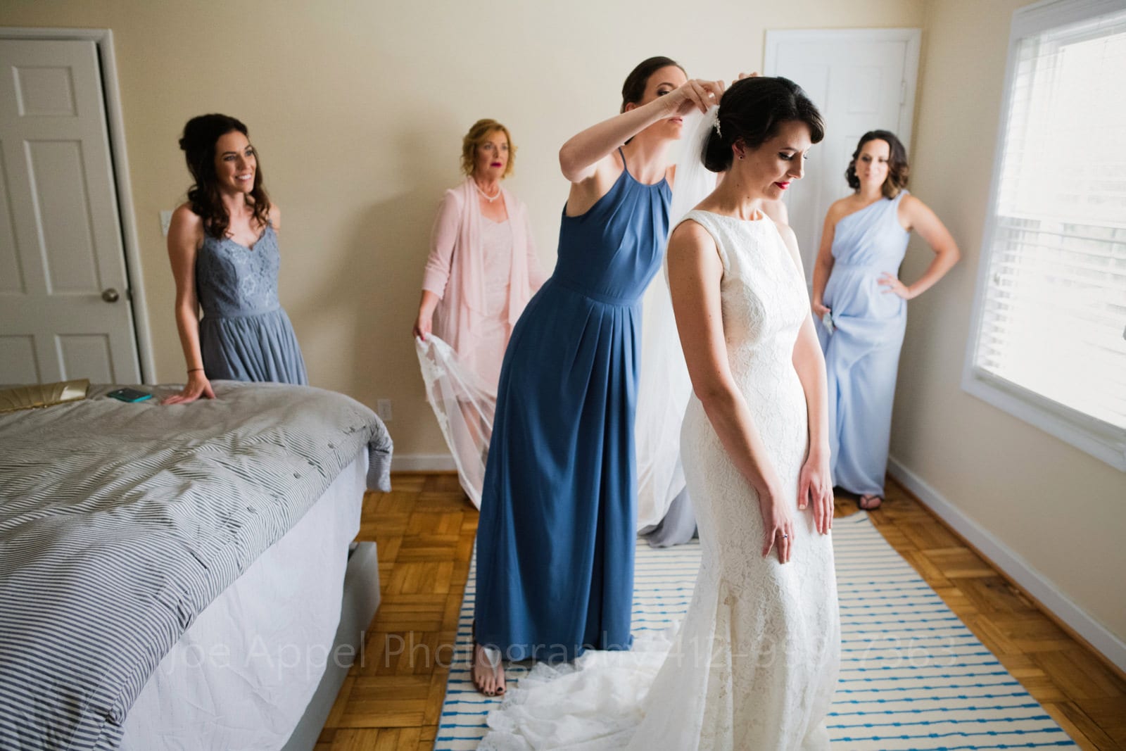 A bride wearing a white gown has her veil put in by a bridesmaid in a blue dress while other bridesmaids wearing blue dresses and her mother in a pink dress look on.