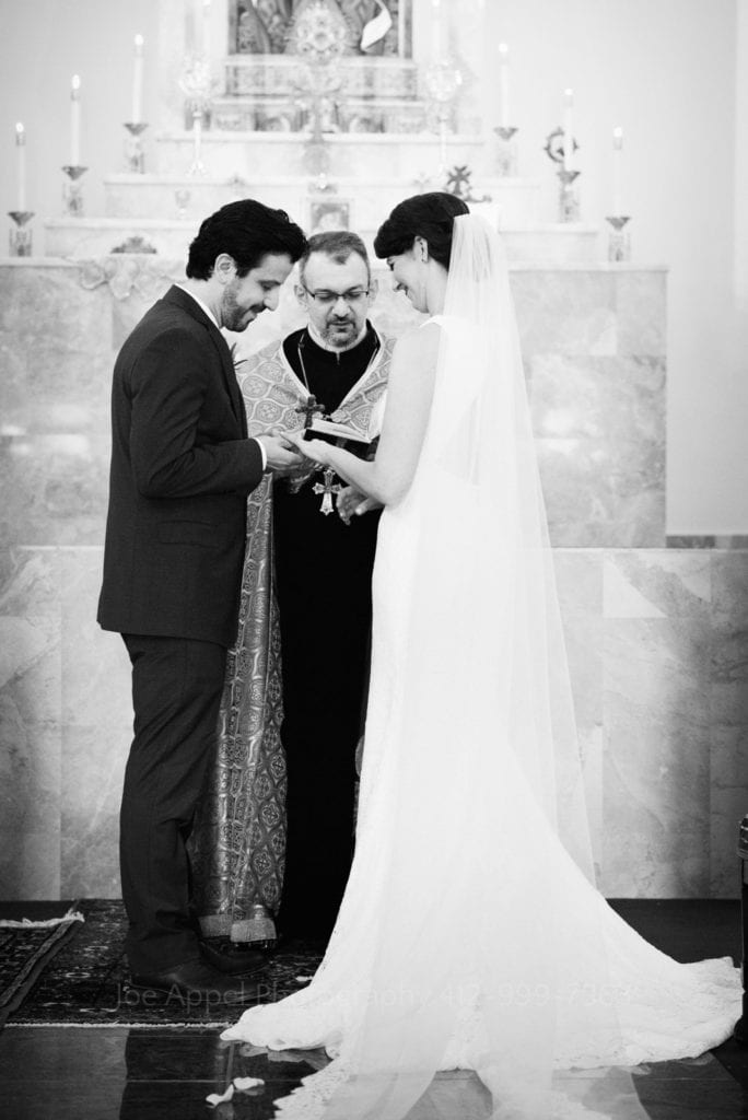 A bearded groom places a wedding band on a bride in a long white dress in front of a priest at St. Mary's Armenian Apostolic Church in Washington DC.