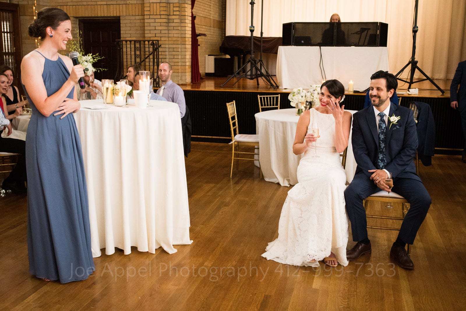 A seated bride wipes a tear as she and her groom listen to a toast from her blue dress wearing matron of honor during their St Francis Hall Washington DC wedding.
