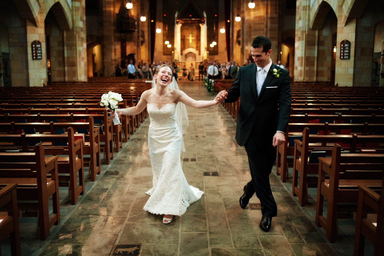 A bride holding a bouquet in one hand and her groom's hand in the other looks up and laughs as they walk down the aisle at the end of their wedding at Sacred Heart Church in Pittsburgh.