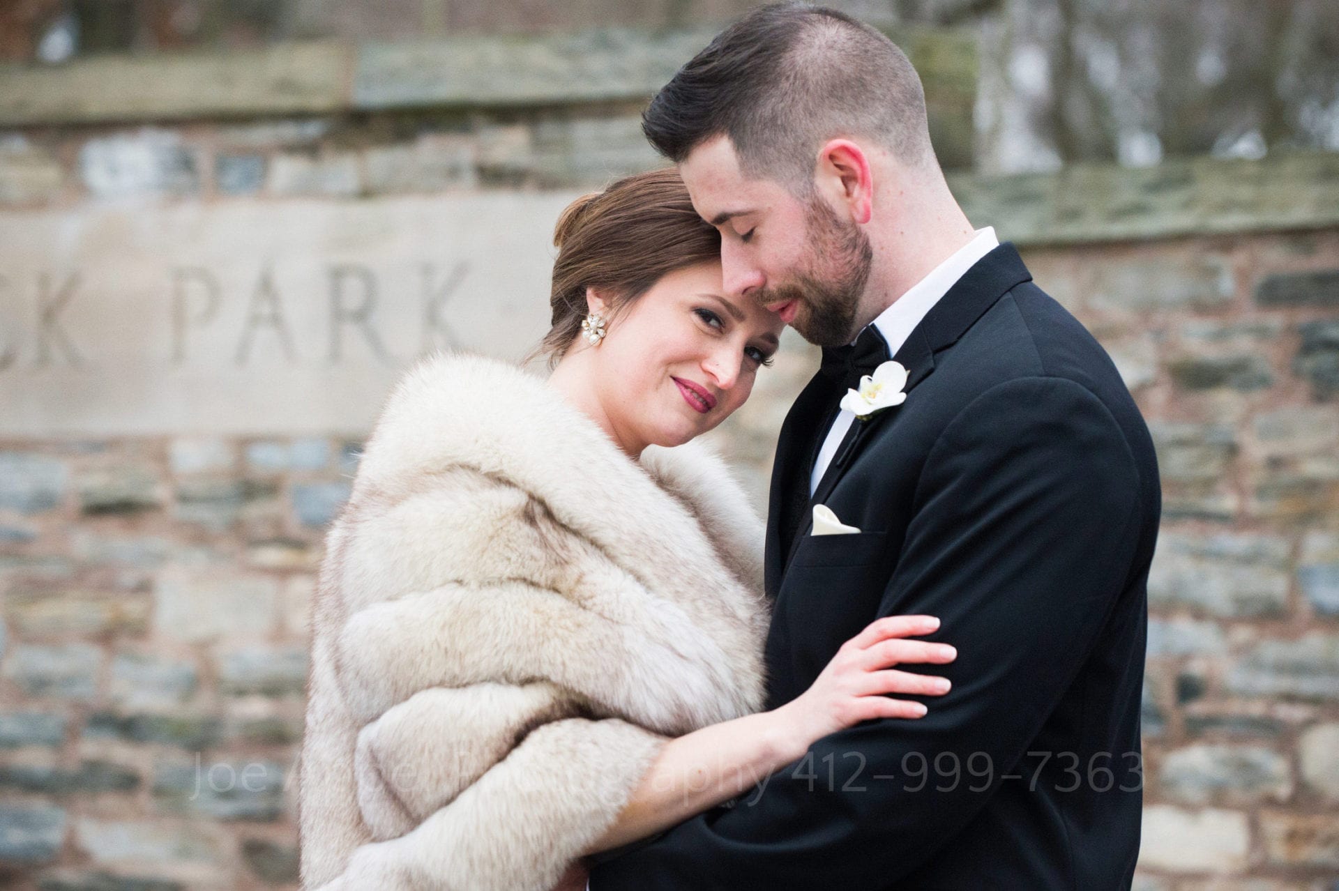 A young couple embraces in front of a stone wall. She wears a fur stole and looks at the camera while leaning into his chest. He wears a black tuxedo and has his eyes closed as his forehead touches hers.