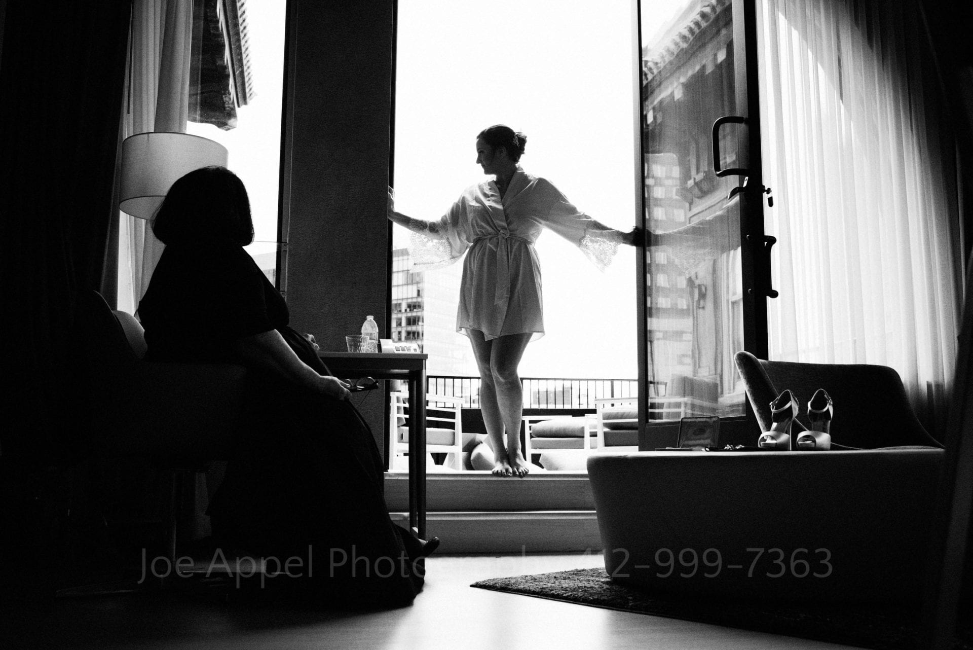 A woman wearing a dressing gown stands in silhouette in a doorway leading to a deck outside of a hotel suite.