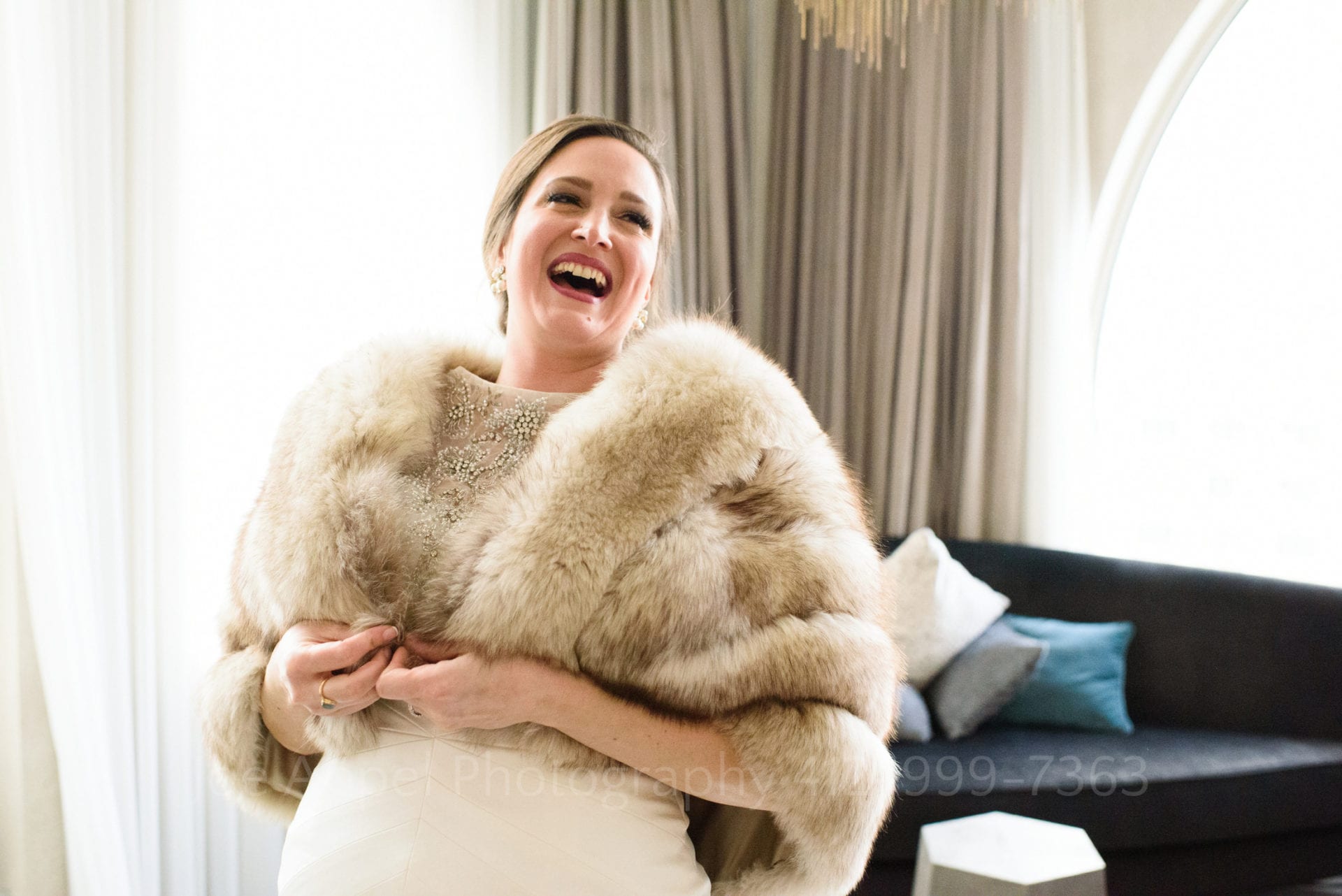 A bride laughs as she buttons her fur stole while standing in a brightly lit hotel room.