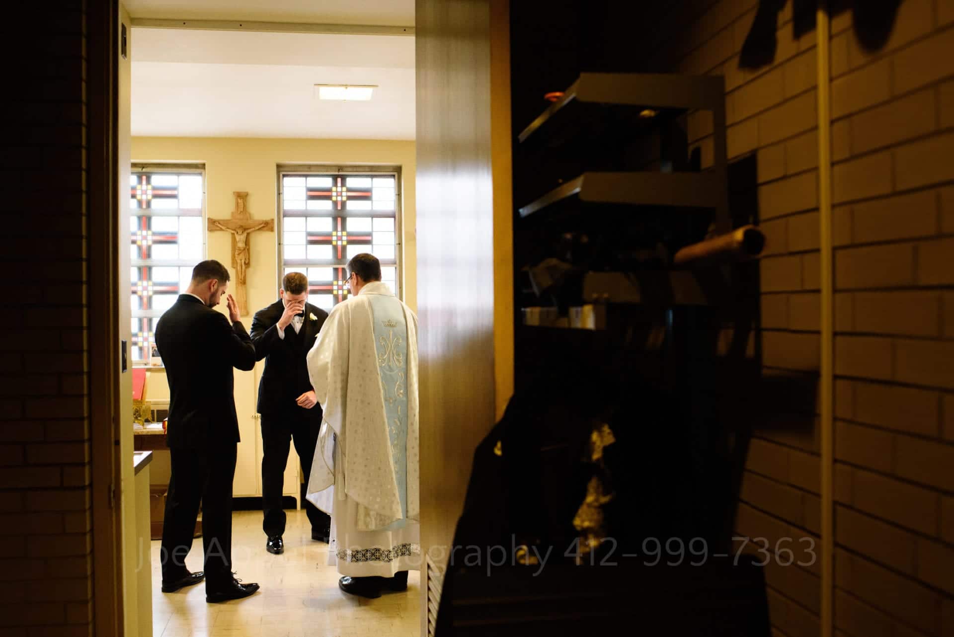 Seen through a doorway, a groom and his twin brother cross themselves while saying a prayer with a priest in white and light blue vestments prior to the groom's wedding ceremony.