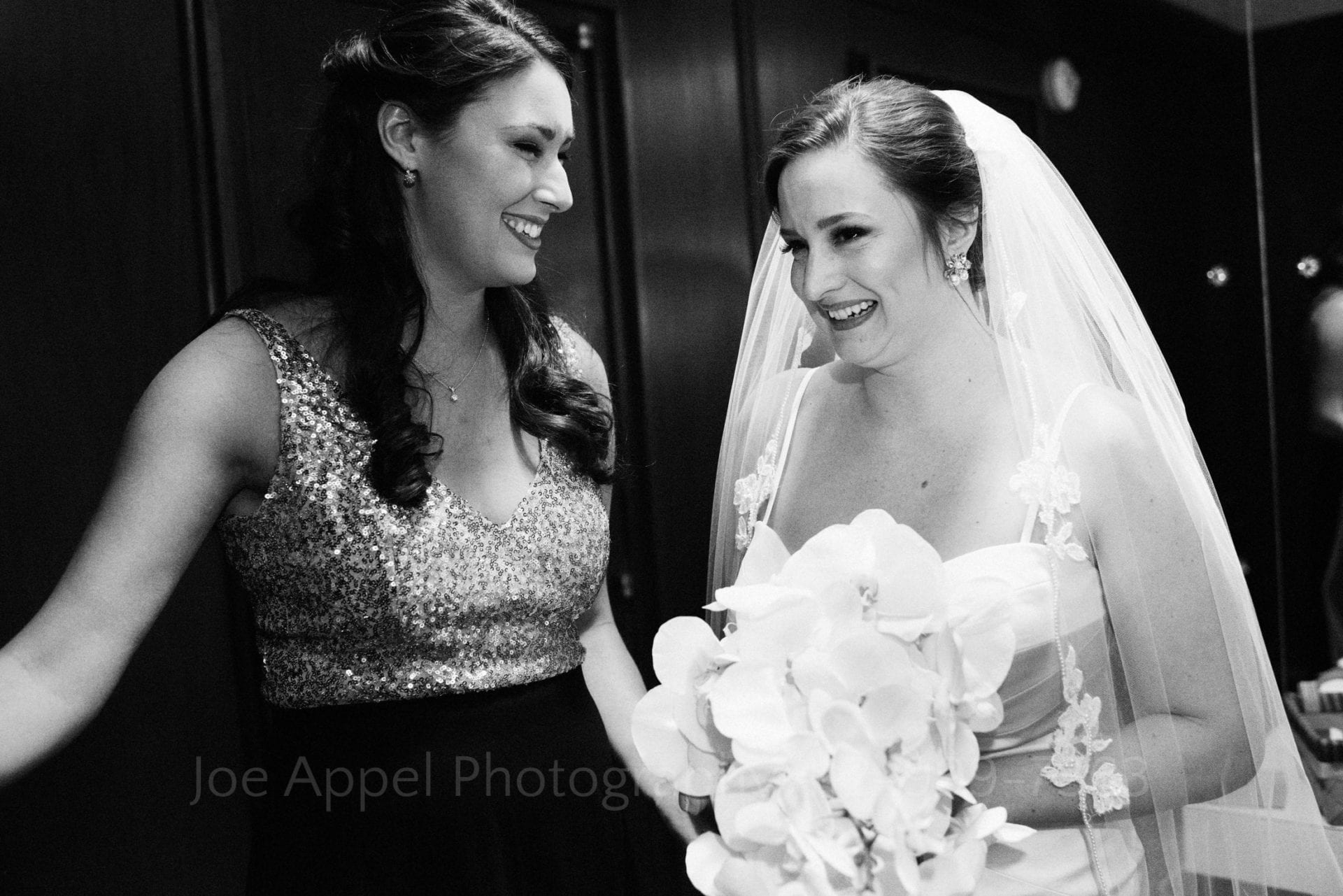 A bride holds her bouquet as she stands in a dark room with her bridesmaid who is wearing a dress with a sequined top.