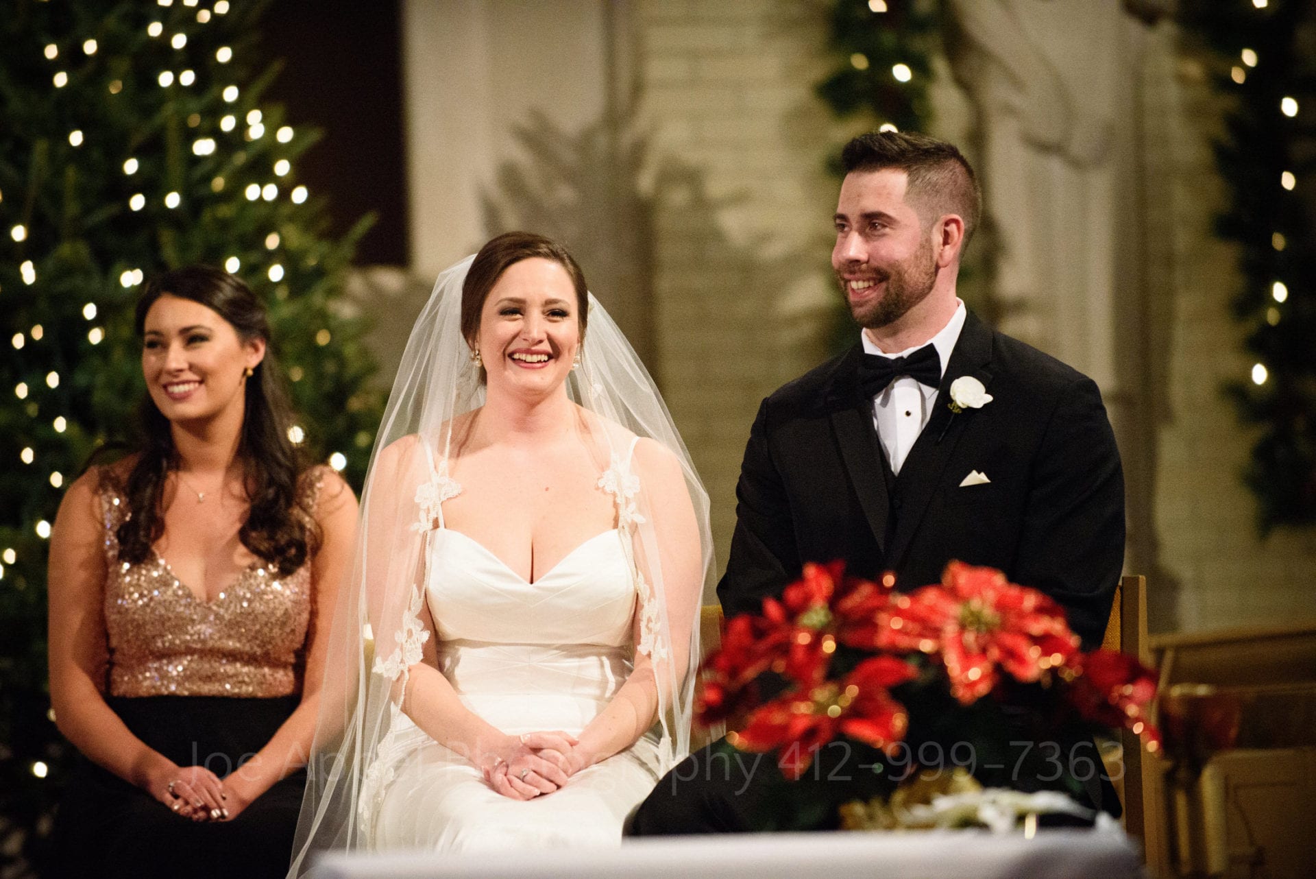 Bride and groom smile as they sit together on an altar. Lighted Christmas trees are behind them and a poinsettia is in the foreground. The maid of honor sits to the left of the bride.