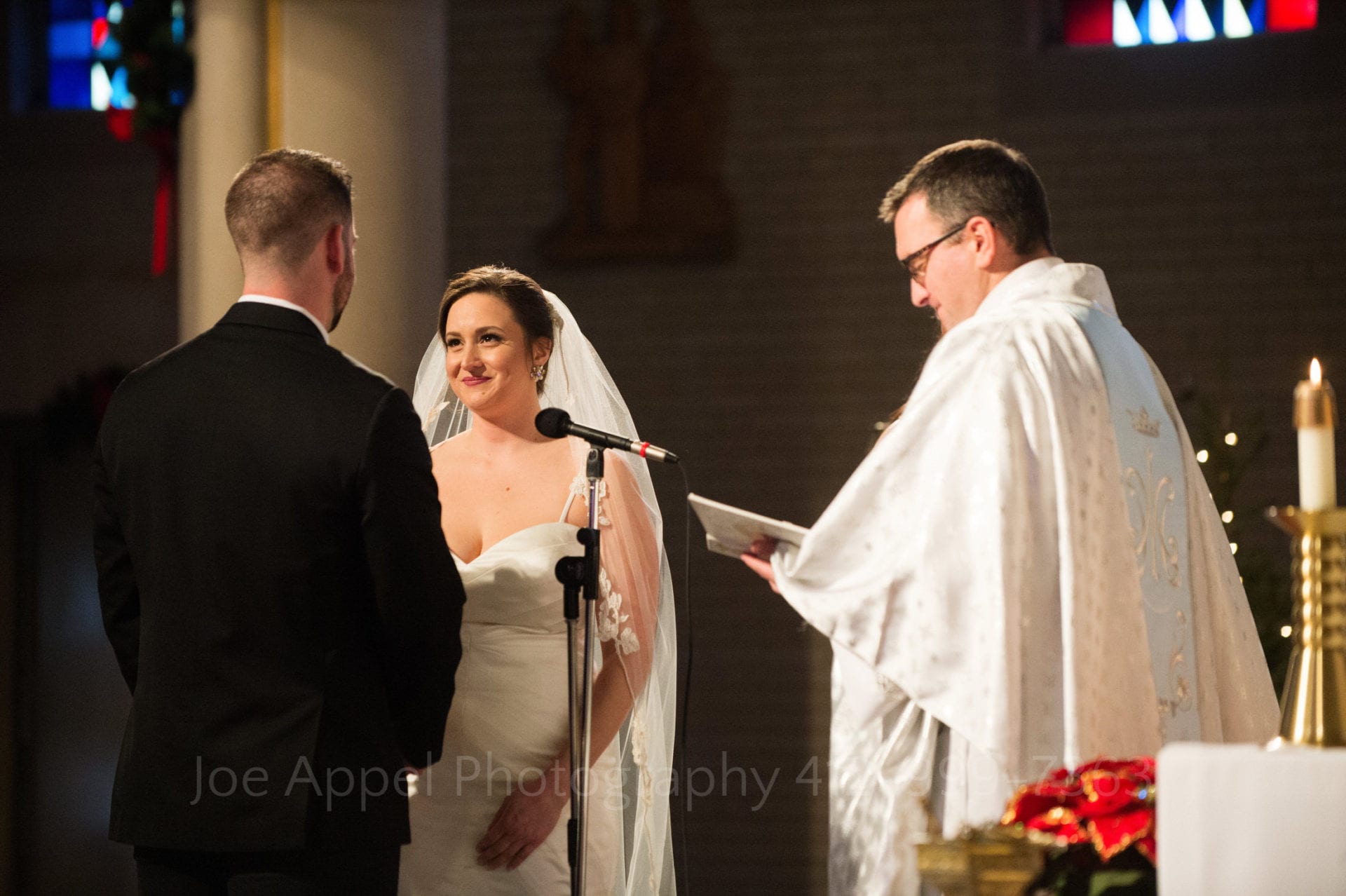 As the priest on the right side of the frame reads from a folder, a bride looks at her groom (his back to the camera) as she holds his hands while exchanging vows.