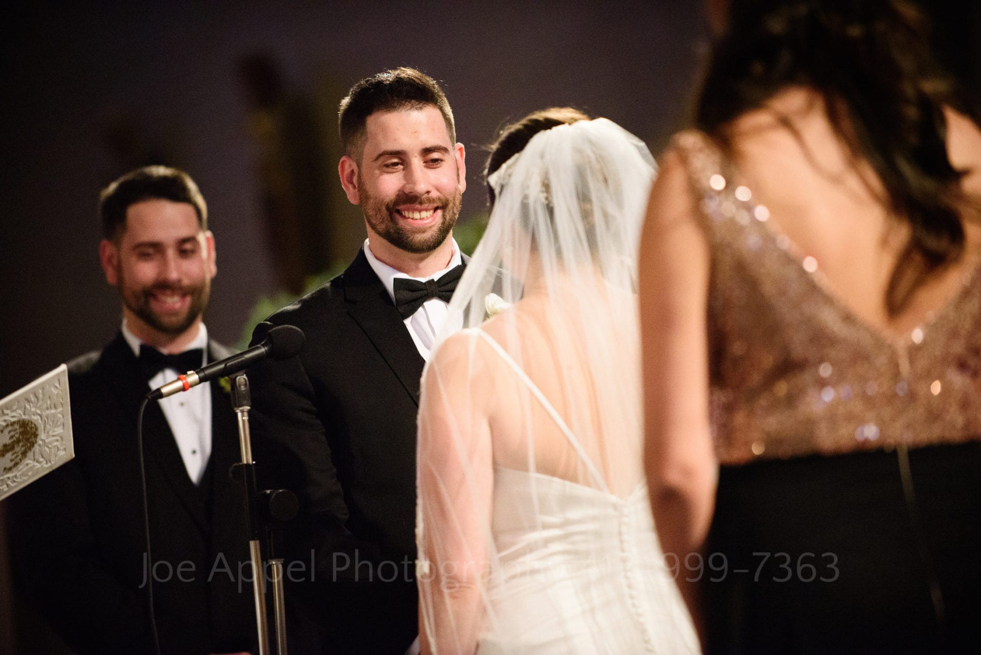 A groom smiles at his bride as they exchange vows. The groom's twin stands behind him.