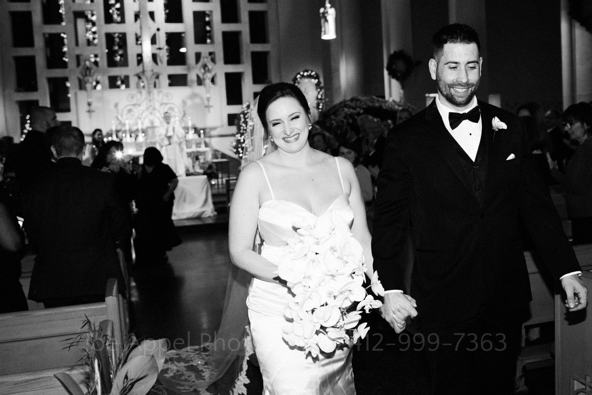 Bride and groom smile and hold hands as they walk down the aisle of St. Raphael parish as man and wife.