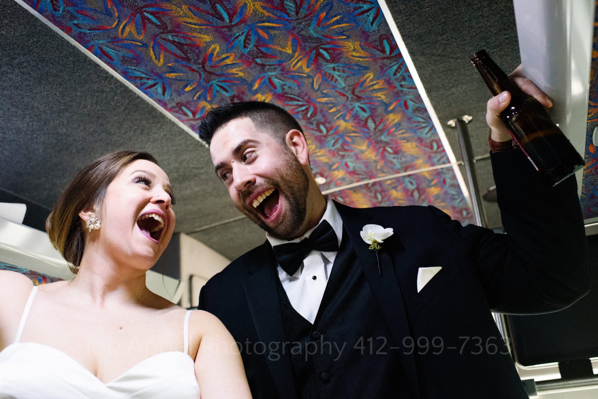 Seen from below, a bride and groom sing to each other as they ride in a limousine bus. The groom holds a bottle of beer in his left hand.