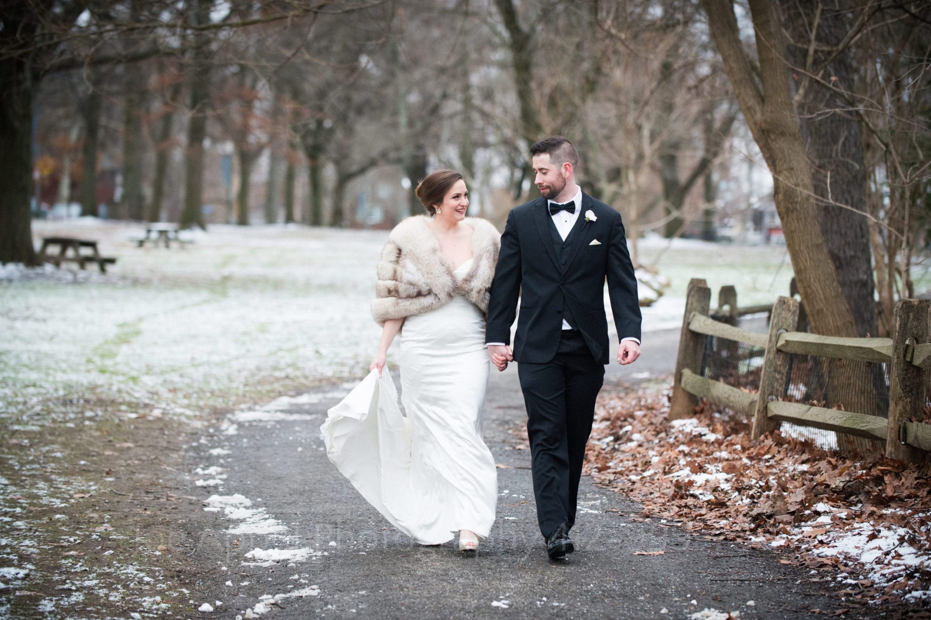 A bride and groom hold hands and look at each other as they walk down a gravel path next to a split rail fence on one side and a snow dusted forest glade on the other in Frick Park.