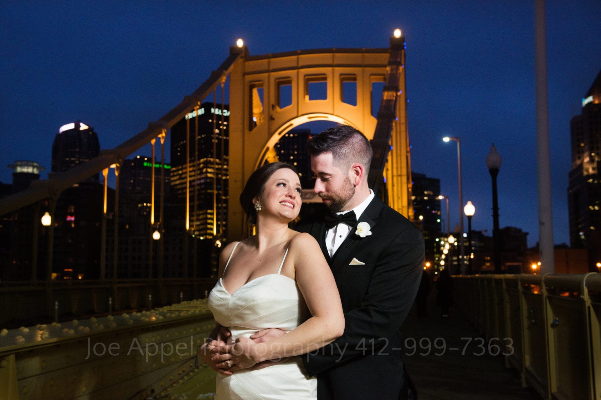 A bride looks back at her groom as he holds her from behind in front of the Roberto Clemente Bridge. The skyline of downtown Pittsburgh is behind the bridge.