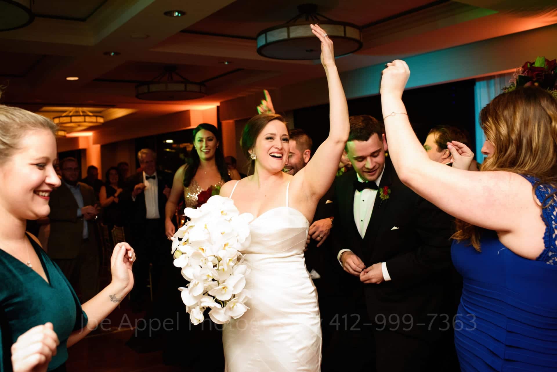 A bride holds her left arm into the air and smiles as she walks through a crowd of guests at her wedding.
