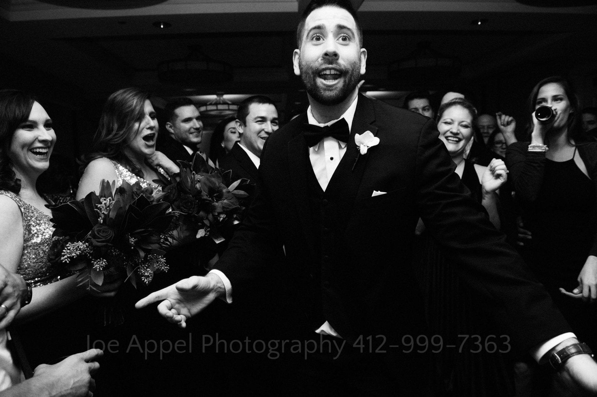 A groom dances in the foreground of the frame while guests surround him.