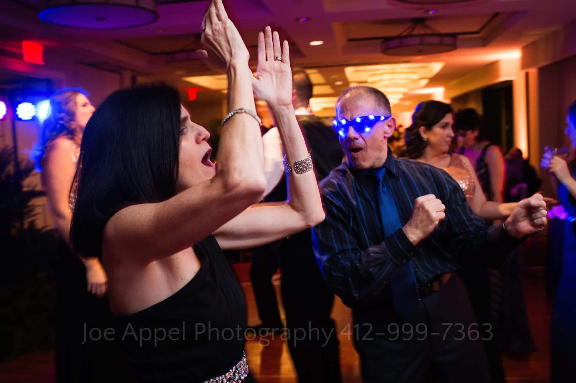 A woman yells and claps her hands in the air as she dances with a man who wears blue sunglasses that have LED lights on them.