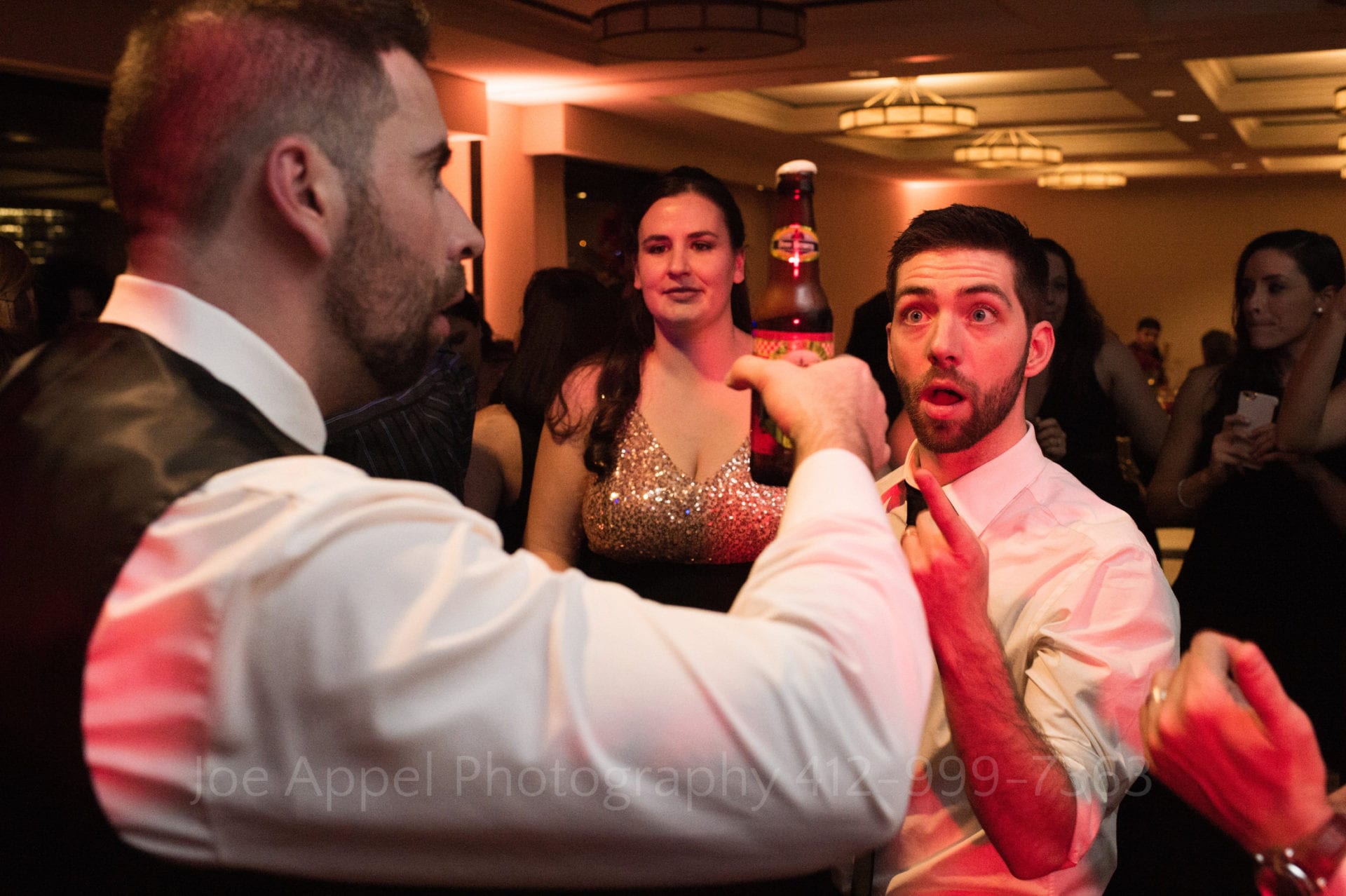 A beer foams over in a grooms hand while a guest with a surprised look on his face points at it.