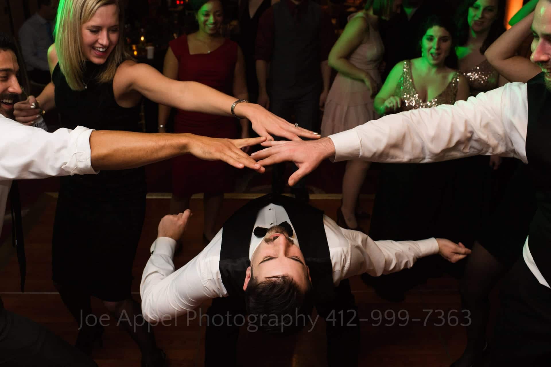 Three people hold their hands out as a man wearing a black vest and bow tie limbo dances beneath them.