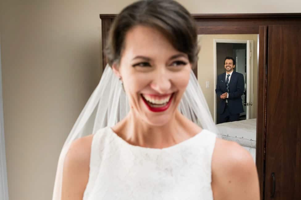 A bride smiles broadly as she sees her groom walk into a room. The groom, seen in the mirror behind the bride, smiles and rubs his hands together.