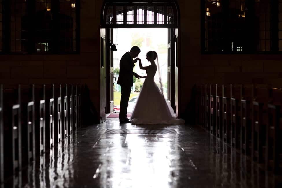 A bride and groom are silhouetted in the doorway of Sacred Heart Church in Shadyside. The groom holds one of the bride's hands up to kiss it.