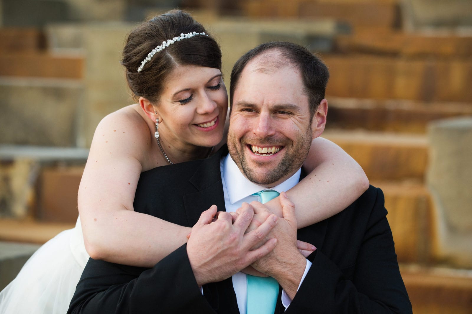 A bride with a beaded hairband holds her arms around the shoulders of her groom who is wearing a light green tie and a black suit.