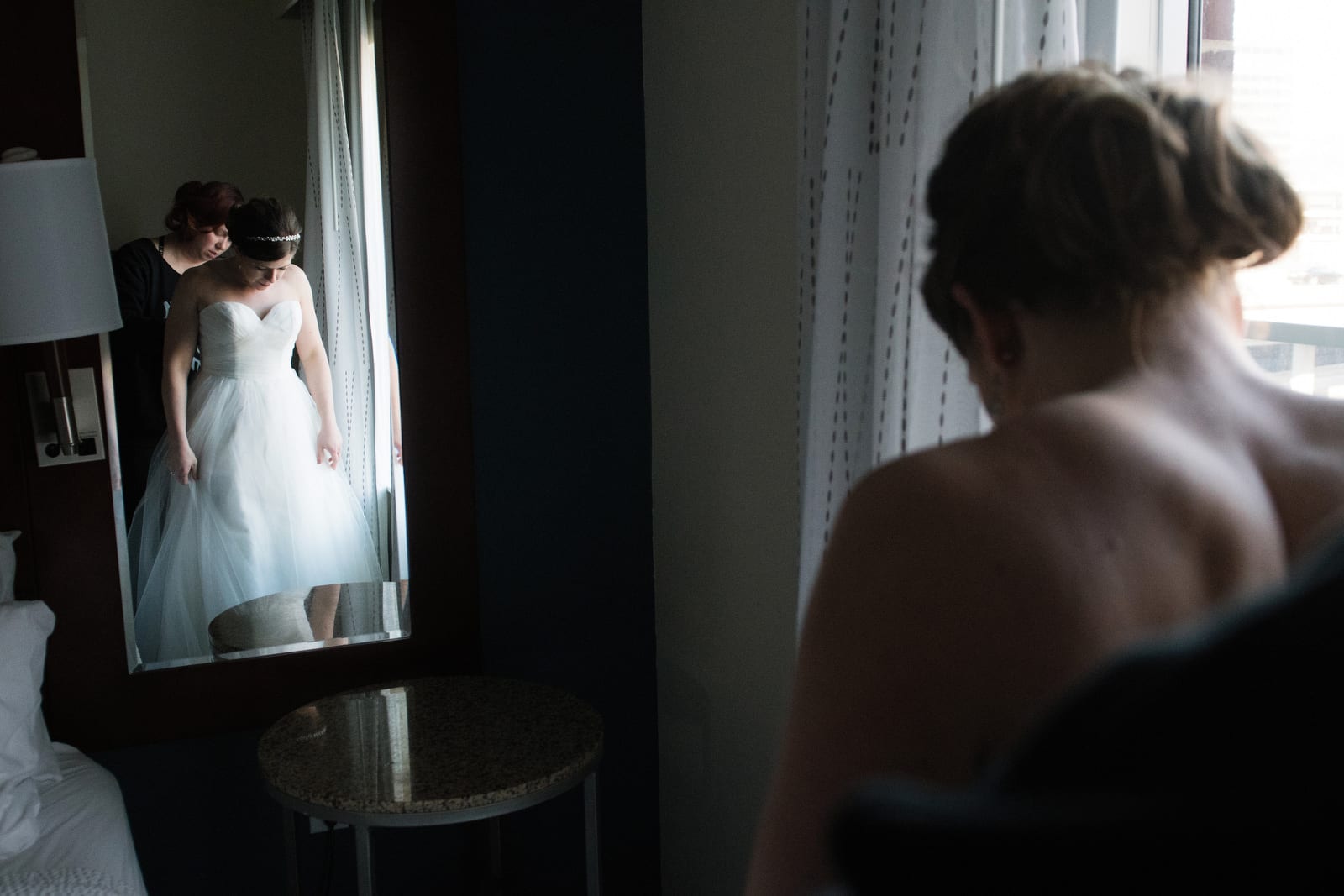 A bride is reflected in a floor length mirror as her friend stands behind her and buttons up her dress.