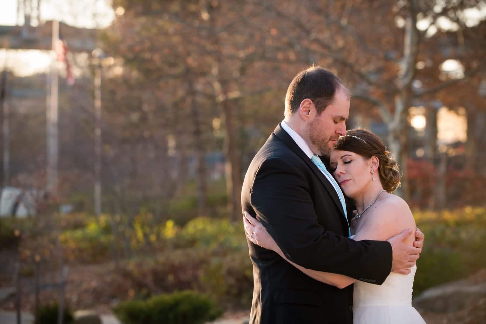 A bride and groom close their eyes and hug with the warm afternoon sunlight behind them.