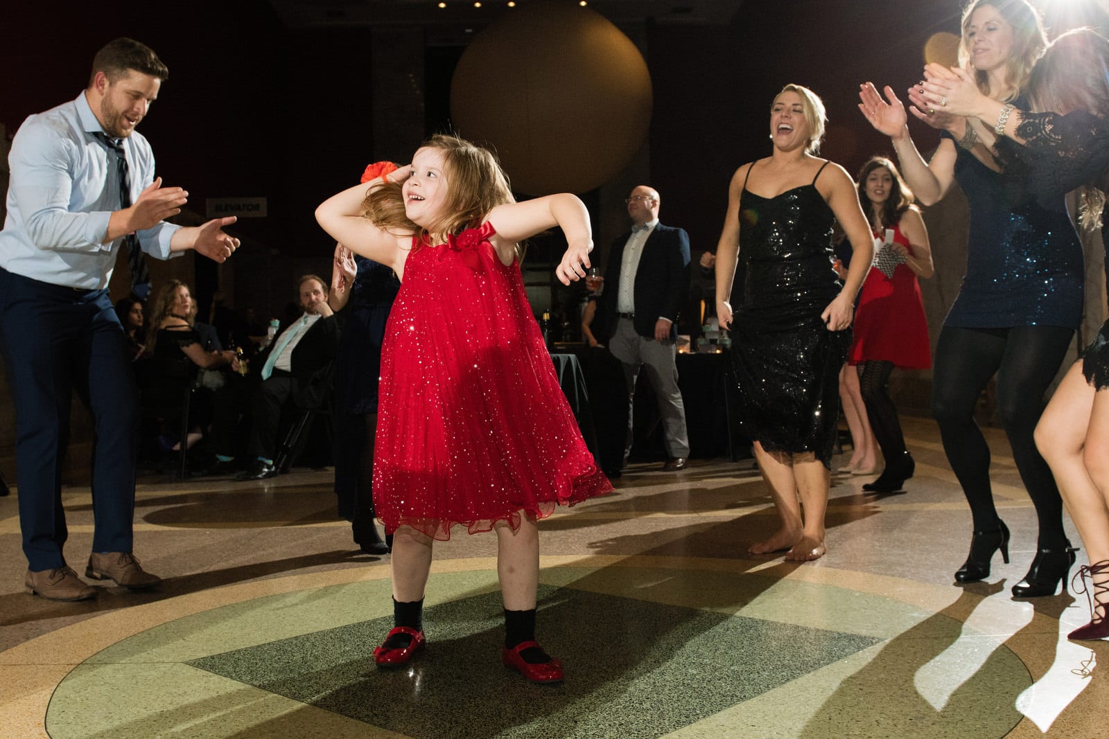 A little girl in a red dress dances as adults around her clap their hands during a wedding at the Pittsburgh Children's Museum.