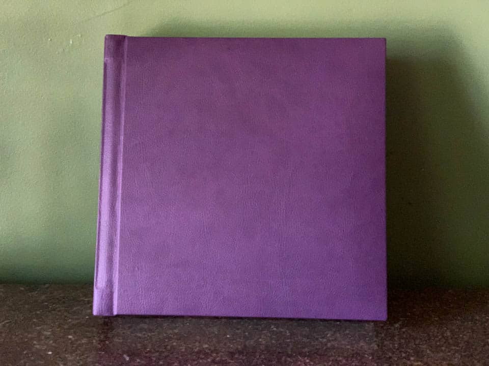 A purple leather covered wedding album leans against a green wall.