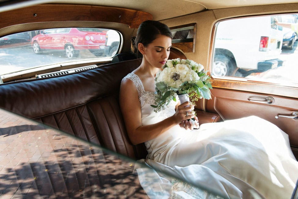 A bride seated in the back of an antique limousine sniffs the flowers of her bouquet.