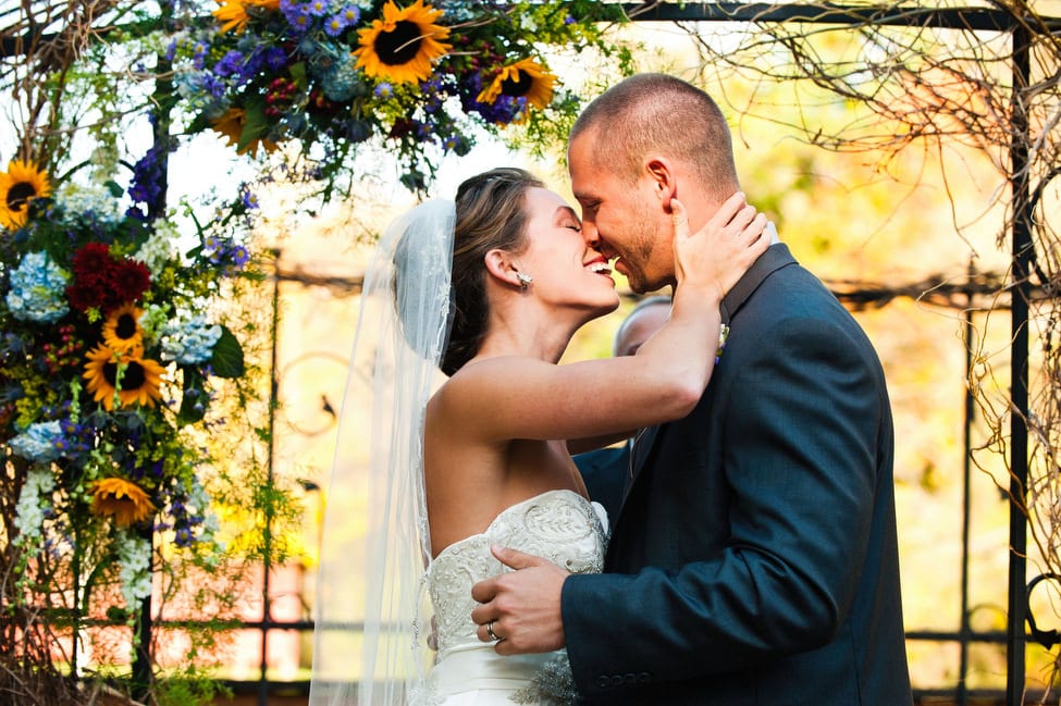 A bride and groom kiss in front of a flowered arbor at an outdoor ceremony.