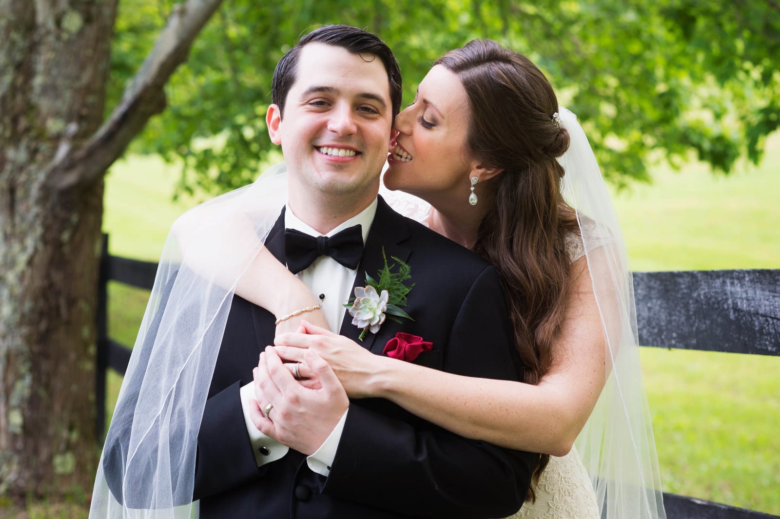 A bride and groom stand in a field next to a split rail fence. She hugs him from behind and bites his earlobe as he smiles.