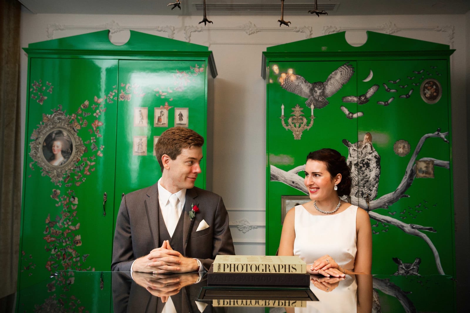 A couple sits at a table at the Hotel Monaco Pittsburgh in front of green painted cabinets with owl decoupage decorations. The couple looks at each other and smiles.