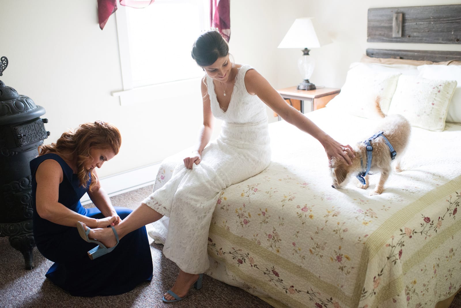 A bride pats the head of her small dog as she sits on a bed. A bridesmaid in a blue dress kneels on the floor putting the bride's shoes on.