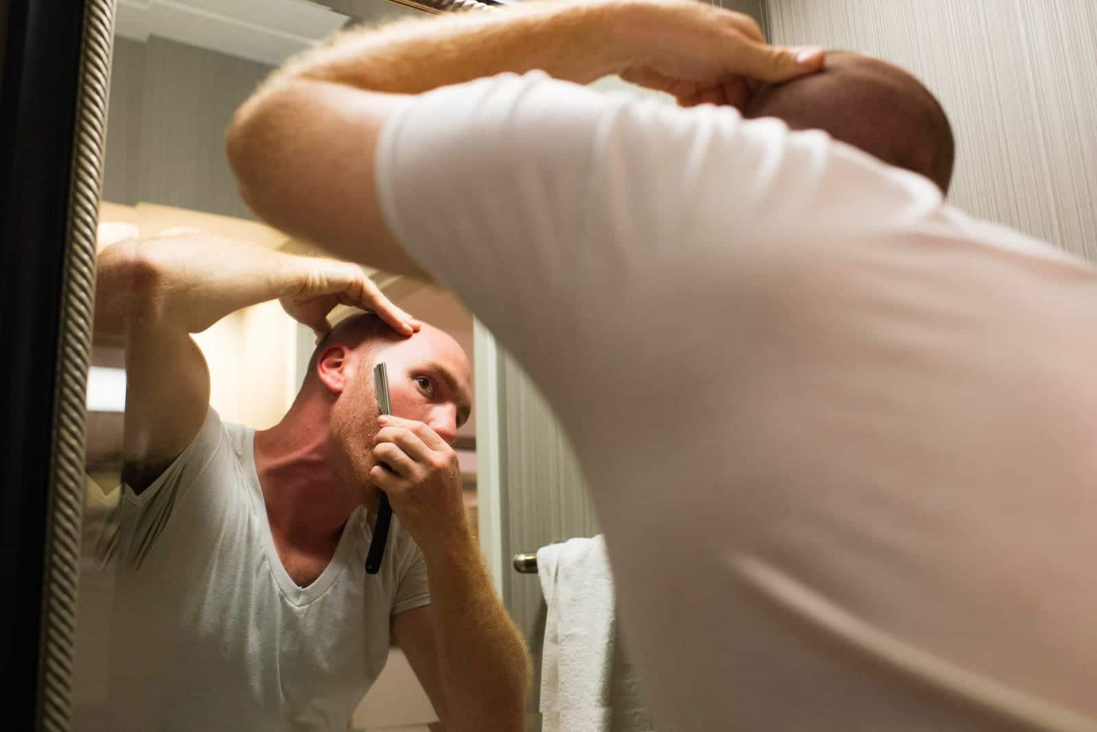 A man looks into a mirror as he uses a straight razor to shave his beard.