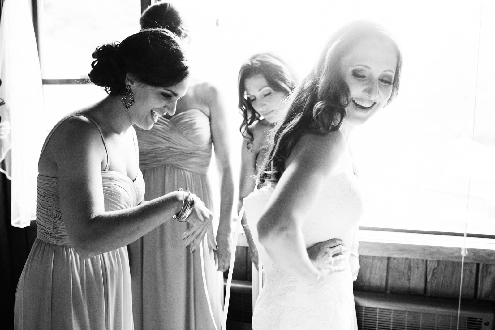 A bride looks over her shoulder and smiles as her bridesmaids stand behind her and laugh as they try to figure out how to lace the dress up.
