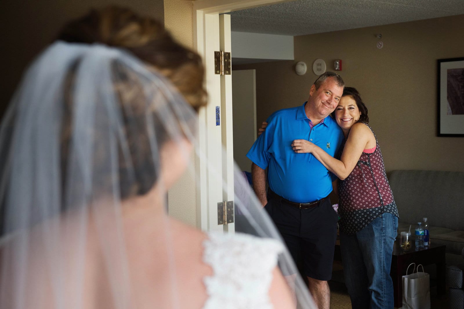 A bride seen from behind as her smiling parents embrace and look at her in her wedding dress.