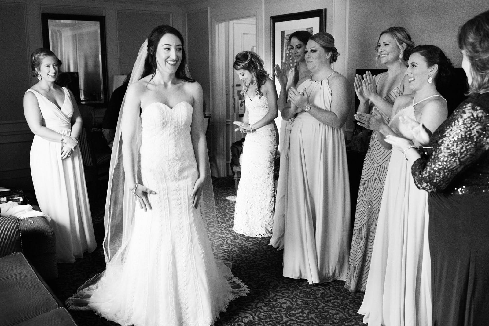 A smiling bride stands in front of her bridesmaids as they applaud in a room at the Omni William Penn Hotel.