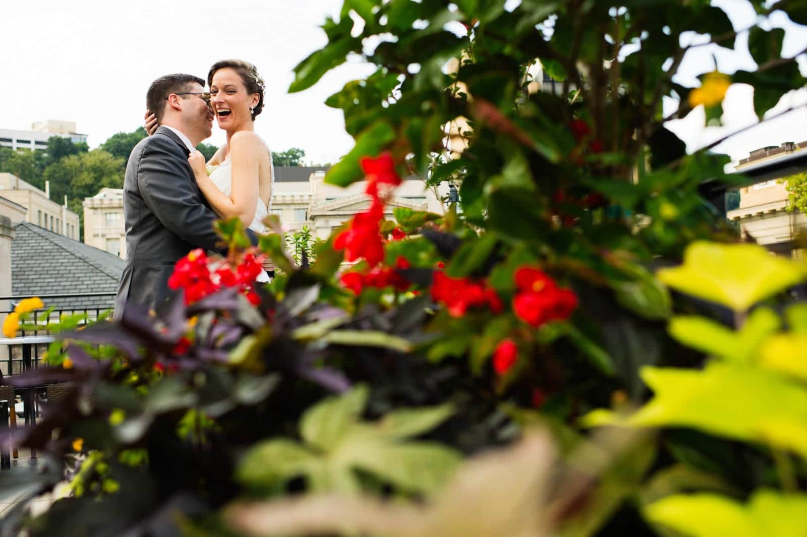 A bride looks at the camera and laughs as her groom whispers in her ear. They're seen through a plant with red flowers as they stand on the rooftop of the University Club.