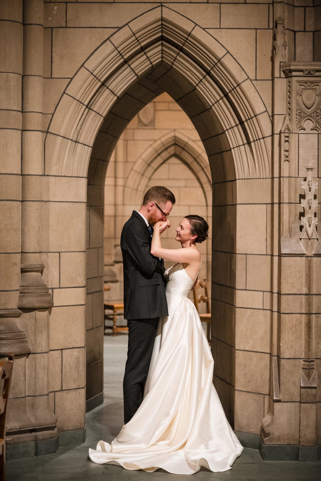 A bride and groom stand in an archway inside of the Cathedral of Learning at the University of Pittsburgh. The groom holds the bride's hand up and kisses it.