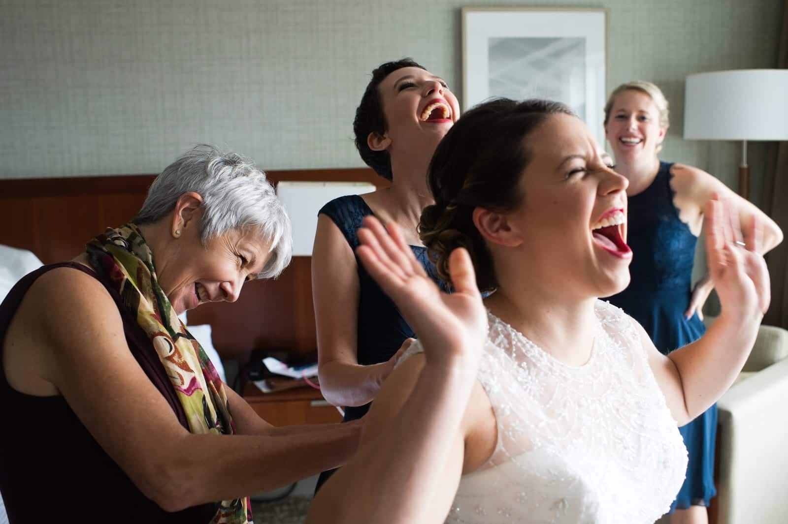 A bride holds her hands in the air and laughs as her mother buttons up her wedding dress. The mother laughs too, as do the two bridesmaids behind them.